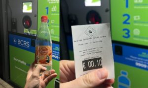 One hand is holding a plastic bottle and one hand is holding receipt with reverse vending machines in the background.