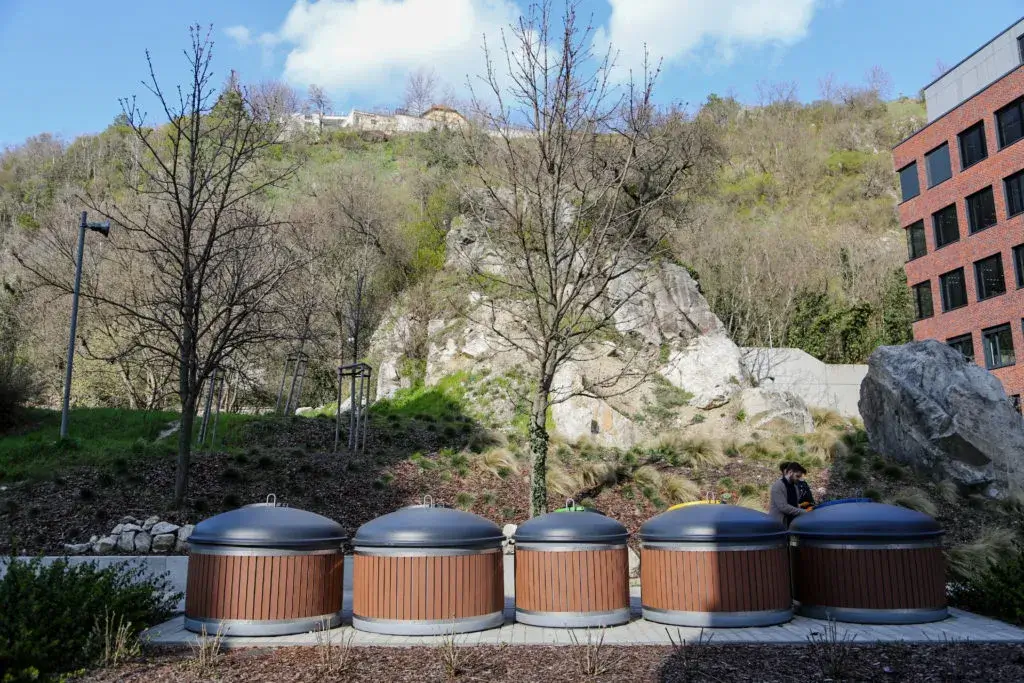 Five semi-underground bins at a garbage stand with urban greenery in background. 
