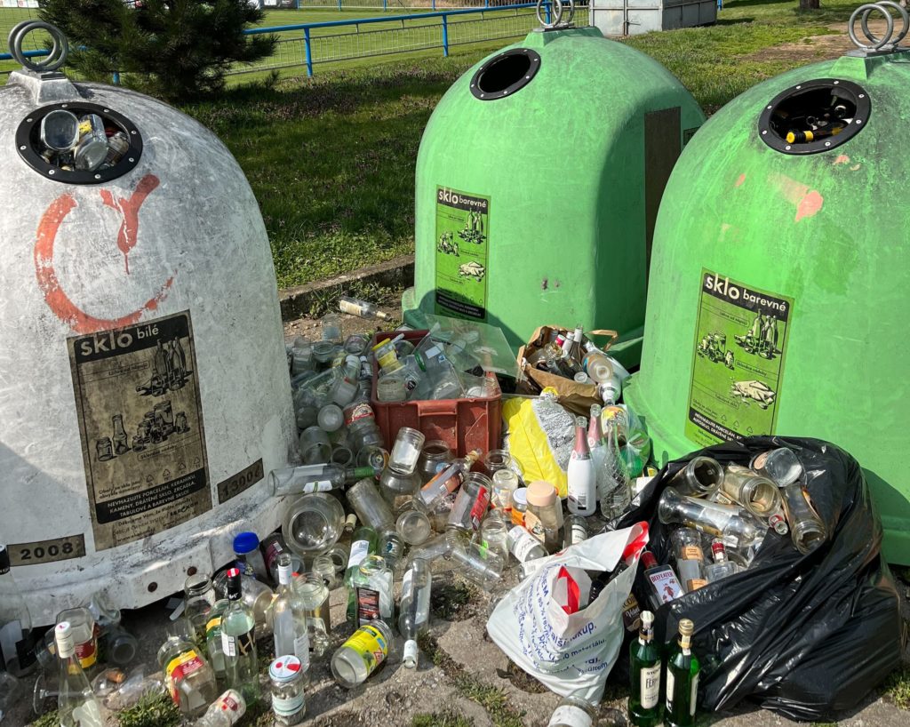 Overflowing glass waste bins with many glass bottles lying next to the bins in the street.
