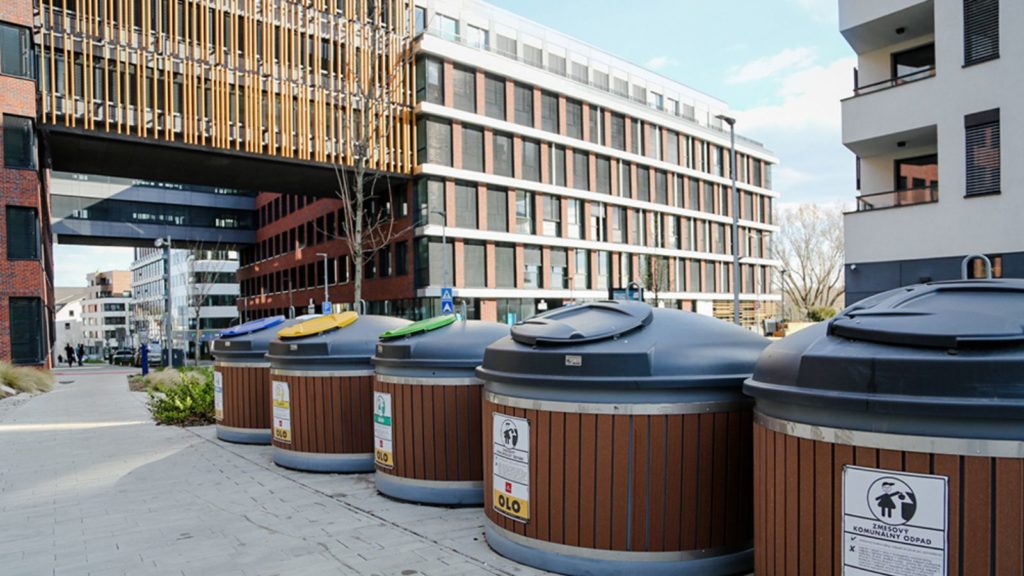 Semi-underground bins for different streams like plastic, paper, and solid waste in front of modern infrastructure and buildings.
