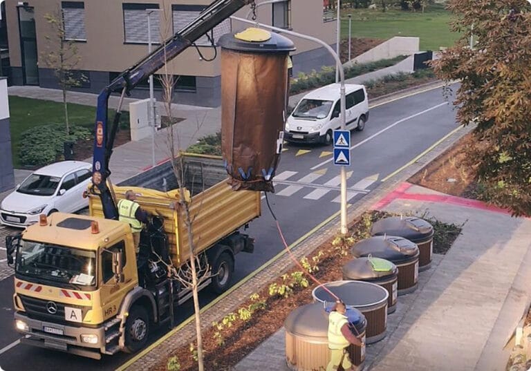A waste collection truck with a crane lifting a waste container, while a worker stands by.
