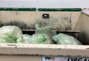 A dumpster with a sensor attatched to its back wall filled with green trash bags.