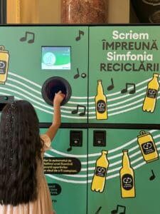 A girl deposits a bottle into a bright green recycling machine decorated with musical notes.