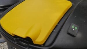 A large outdoor waste container with a yellow lid and a small sensor device attached.