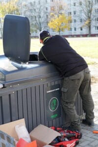 A worker installs waste acess control devices onto a large outdoor waste container.