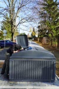 A worker installs wasre access control devices on a large outdoor waste container beside a residential street.