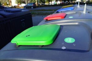 Row of large outdoor recycling containers with coloured lids and small access-control sensors installed.