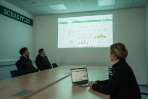 Three colleagues review a projected data dashboard in a meeting room using laptops and charts.