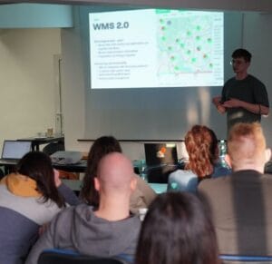 A presenter explains “WMS 2.0” to a small audience, with slides showing a mapped interface.