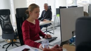 A focused person works at a computer in a bright office with colleagues nearby.