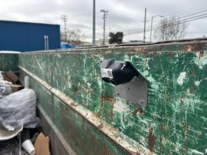 A mounted fill-level sensor attached to a worn, rusty green industrial dumpster wall.