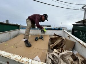 A worker inside a dumpster installs a small sensor device on the inner wall using tools.