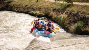 A group paddles a blue raft through fast-moving whitewater on a sunny day.