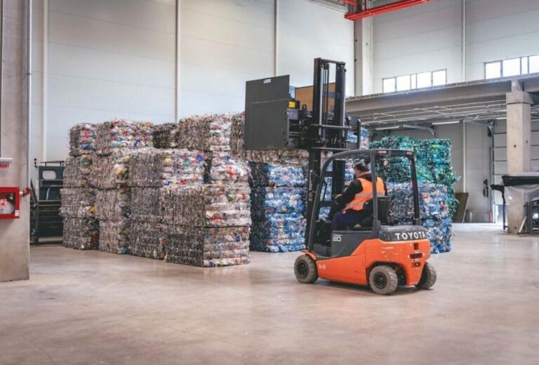 A forklift moves large compressed bales of recyclable plastic inside a warehouse.