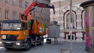 Orange waste-collection truck using a crane to lift an underground container in a city street.