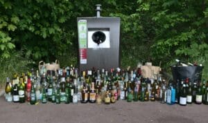 Dozens of glass bottles piled around an overflowing glass-recycling container in a leafy area.