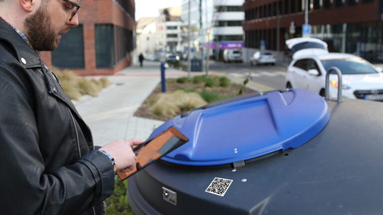 Person scanning a QR code on a large outdoor bin using a tablet.
