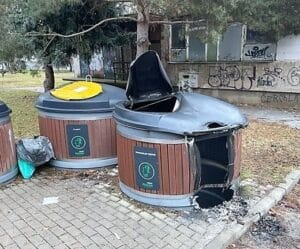Burned and melted semi-underground waste container heavily damaged beside intact neighboring bins.