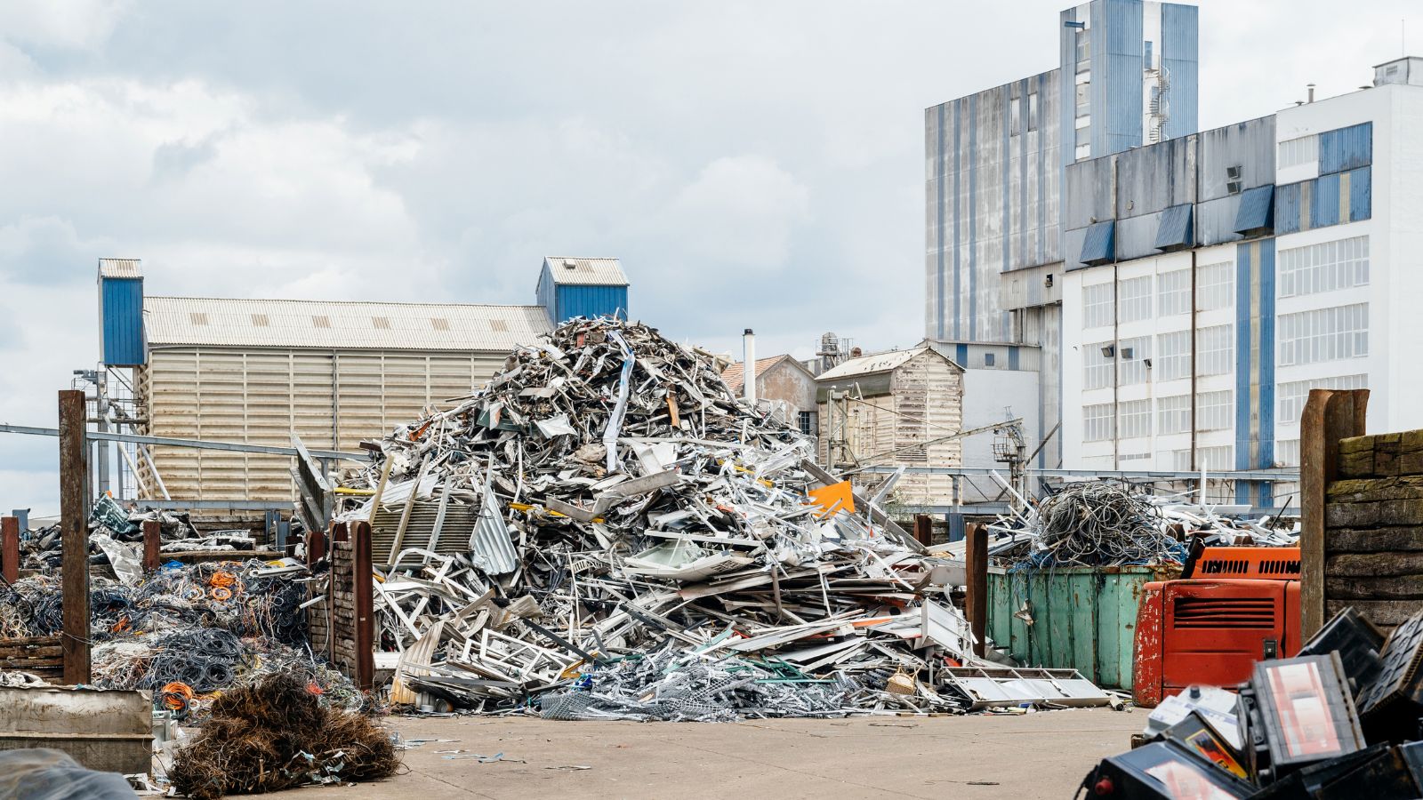 A pile of metal scrap and wires in an industrial recycling yard, surrounded by factory buildings.