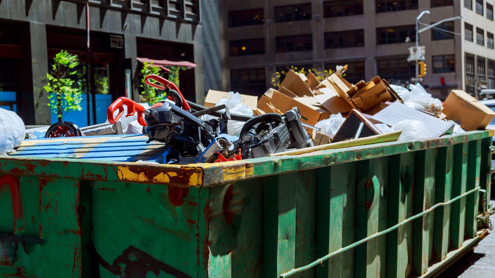 An overfilled dumpster with cardboard boxes, plastic waste, and discarded tools in an urban setting.