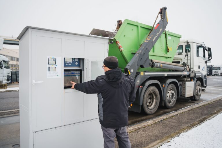 Worker using an outdoor control terminal while a truck lifts a large green container nearby.
